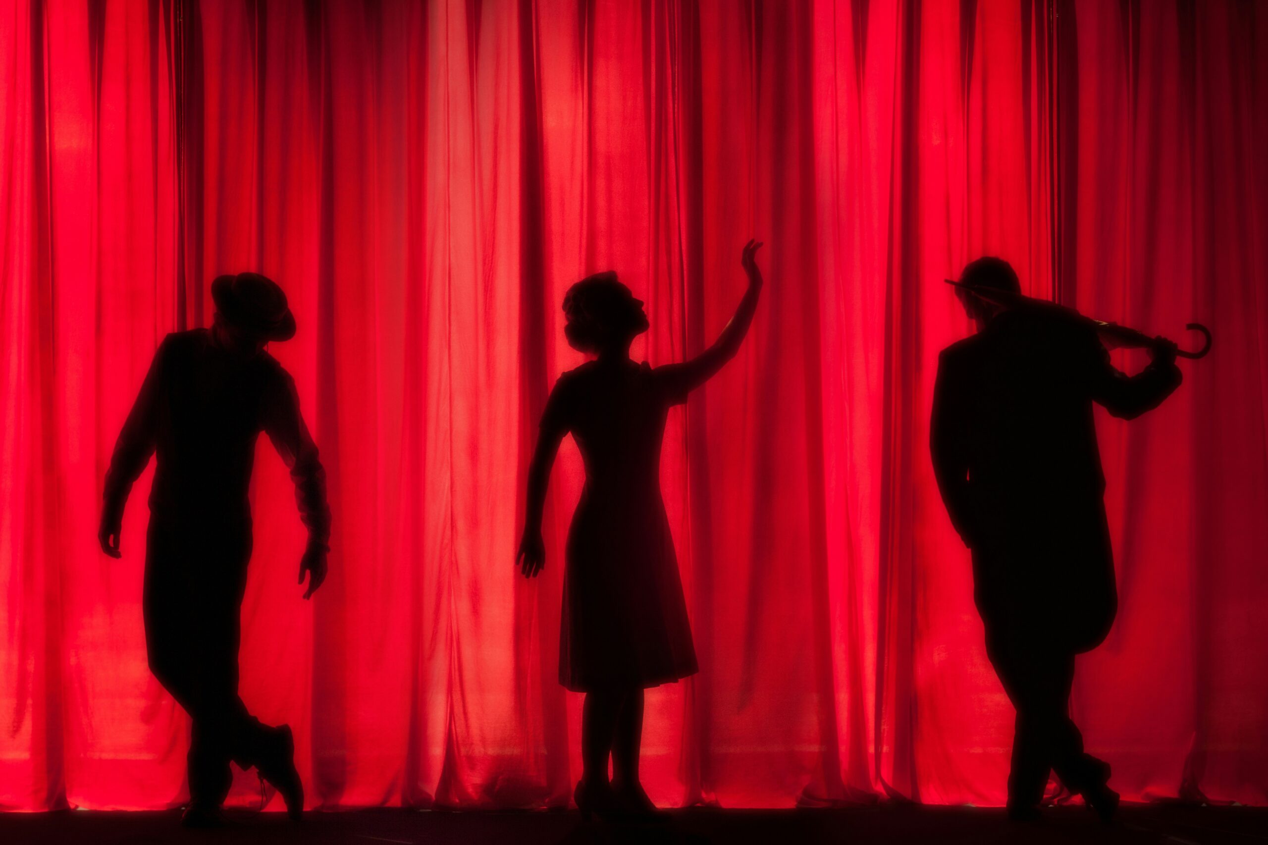 Three silhouetted performers posing in front of a brightly lit red stage curtain.